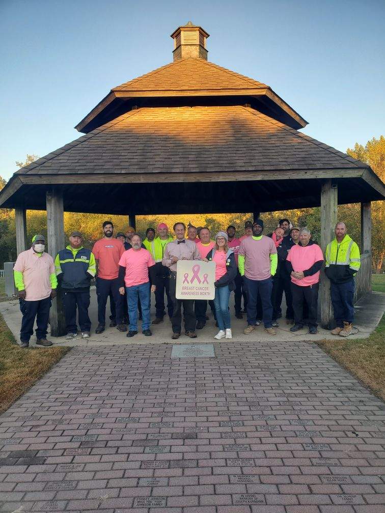 Volunteers pose under an outdoor pavilion, wearing pink shirts, with a breast cancer awareness banner.